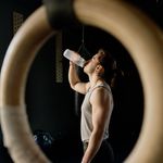 Caucasian man drinking water in a gym setting, framed by gymnastic rings.
